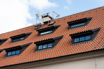 Red clay tiled roof featuring six black-framed dormer windows and rooftop antennas under a blue sky. Suitable for architecture, construction, and real estate design reference or educational use.