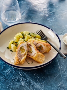 Juicy chicken kiev, breaded and golden, is plated alongside seasoned potatoes and fresh herbs. The dish is elegantly arranged on a blue table, inviting diners to enjoy a delightful meal