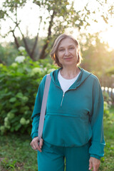 Happy smiling active mature woman in blue sportswear with yoga mat on her shoulder outdoors. Sport training concept