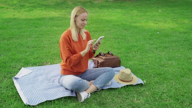 Beautiful young blonde woman sitting on picnic blanket in green park, smiling and happily scrolling through social media on smartphone during relaxing sunny day outdoors.Business,technology, blogging,