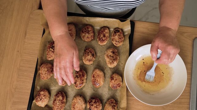 Brushing oil onto baked cutlets on tray. Unrecognizable person greasing golden meat patties. Spreading fat over portions with kitchen brush. Preparing flavorful homemade juicy meat cutlets
