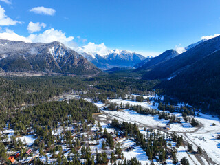 Drone shot looking down a mountain valley featuring dense pine forest, snowy ground, and a small...