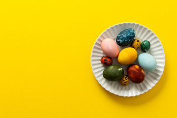 A variety of beautifully painted Easter eggs in different colors and patterns are placed on a delicate white plate. The vibrant hues stand out against the cheerful yellow backdrop