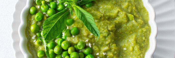 A bowl of vibrant green pea and broccoli soup is topped with fresh mint leaves for flavor. This healthy dish is perfect for a warm lunch or cozy dinner at home, banner