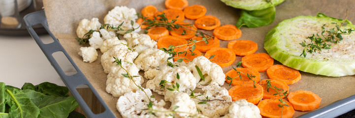 Cauliflower, carrots, and cabbage are sliced and arranged on a tray lined with parchment paper, ready for roasting with herbs in a warm kitchen setting, banner