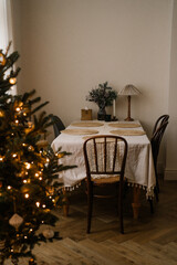 Minimalist Christmas tree decorated with ornaments and glowing fairy lights, next to an elegant dining table prepared for the holidays and Christmas dinner
