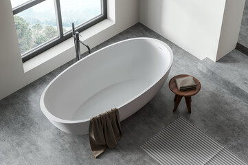 Fototapeta na wymiar White freestanding bathtub in modern bathroom interior, top view, with gray concrete floor, large window and wooden stool. Concept of home relaxation. 3D Rendering