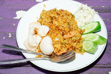 fried rice dish on a white plate with toppings of sliced ​​cabbage, cucumber and some crackers isolated on a purple wooden table surface.