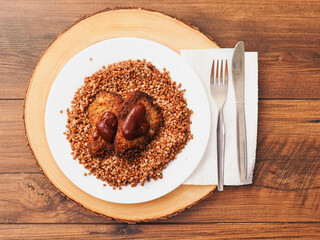 Simple tasty buckwheat and breaded chicken fillet with barbeque sauce meal on a white round plate and round wooden serving board and wooden table. Popular Russian dish..
