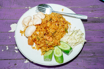 fried rice dish on a white plate with toppings of sliced ​​cabbage, cucumber and some crackers isolated on a purple wooden table surface.