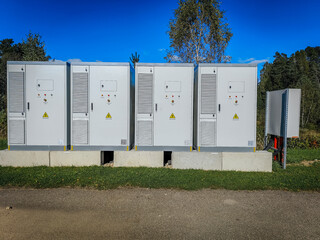 A row of outdoor high-voltage electrical cabinets with warning labels, ventilation panels, and emergency stop buttons under a clear blue sky.