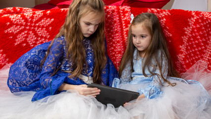 Two sisters in festive dresses sitting in the bedroom decorated with a Christmas tree and a garland, children use a digital tablet computer