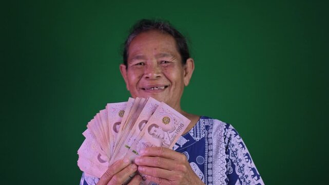 Happy elderly Asian woman holding money Thai baht banknotes with big smile, Financial success, luck, and retirement joy on chroma key green screen background, Grandmother happily with a money