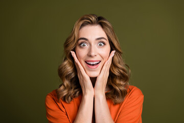 Excited young woman in orange t-shirt posing with hands on face against a green background showing...