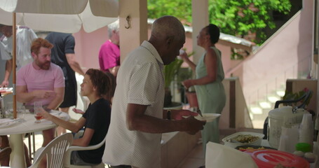 Elderly man serving himself food during an outdoor family gathering, surrounded by diverse people...