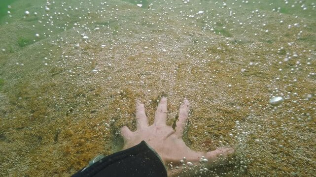 POV shot: Man's hand slides forward along benthic colony of microalgae that synthesizes gas, causes cascade of tiny bubbles to rise upward, fragments of colony disconnected and floats up with bubbles