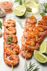 Skewers with tasty grilled shrimps, lime slices, herbs and sauce on white marble table, closeup. Delicious seafood dish