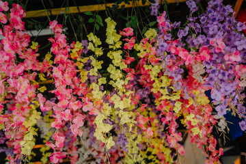 Colorful artificial flowers in shades of pink, yellow, and purple hang in bunches, creating a vibrant decorative display at a market or shop.