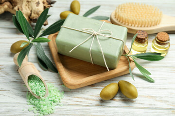 Natural soap bar with olives, sea salt, brush, oil and green leaves on white wooden table, closeup