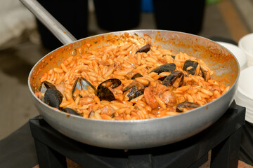 A seafood pasta dish with mussels is being served in a large pan at a vibrant outdoor festival. Event attendees gather around, enjoying the delightful aroma and flavors