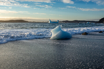 Waves crash around icebergs that have washed up on a black sand beach.