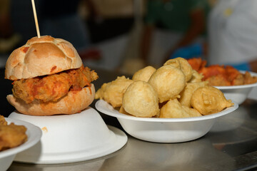 A fried chicken sandwich and bowl of pettole snacks at a bustling food event, several golden-brown fritters are displayed on white plates, hinting at a savory meal, summer food festival in Puglia