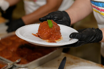 A cook presents a perfectly shaped meatball topped with green basil, accompanied by rich tomato sauce. The dish is part of a community gathering, where attendees enjoy homemade meals