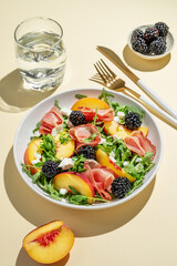 Fresh salad with peaches, arugula, feta, prosciutto and berries in a plate on a yellow background