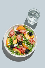 Salad with peaches, arugula, feta, prosciutto and berries in a plate on a blue background
