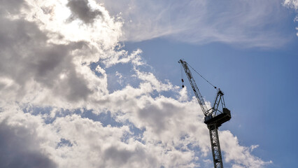 Looking up tall construction crane, sun shines through clouds in background