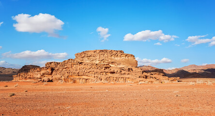 Sun shines to orange rock formations in Wadi Rum desert, few shrubs growing on bright sun lit sand, blue sky with only few clouds above
