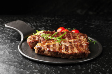 Delicious grilled beef steak with rosemary and tomatoes on dark textured table against black background, closeup