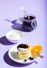 Breakfast of syrniki with sour cream and blackcurrant jam with orange on a lilac background.