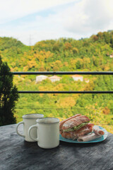 Breakfast in the mountains — two white cups of coffee and a fresh sandwich on a wooden table with a scenic mountain view. Peaceful morning in nature, cozy travel moment.