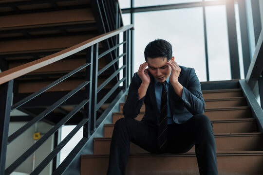 Asian businessman feeling stressed and having a headache, sitting alone on the office staircase, experiencing burnout - Powered by Adobe