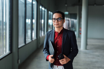 Confident Asian businessman walking in a contemporary office building hallway, holding a folder and...