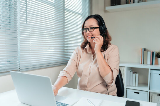 Smiling woman wearing headset with microphone, working remotely on laptop from home office, typing on keyboard