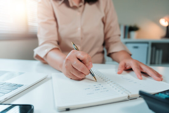 Woman's hand holding a pen, writing financial data and planning notes in an open notebook on a white desk