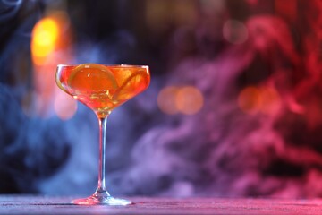 Glass of alcoholic cocktail on table against blurred background with smoke, space for text. Bokeh effect