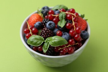 Different ripe berries and basil leaves in bowl on green background, closeup