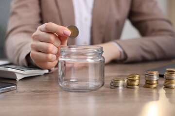 Woman putting coin into glass jar at wooden table indoors, closeup