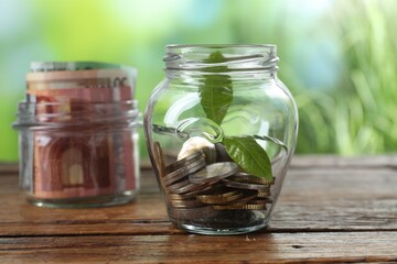 Glass jar with coins and sprout on wooden table against blurred background, closeup