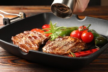 Woman adding pepper to grilled beef steaks, rosemary and vegetables in pan at wooden table, closeup