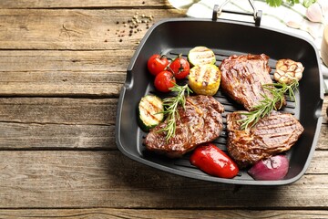 Pan with grilled beef steaks, rosemary and vegetables on wooden table, above view. Space for text