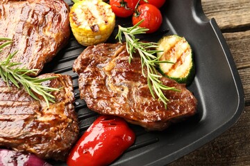 Pan with grilled beef steaks, rosemary and vegetables on wooden table, top view