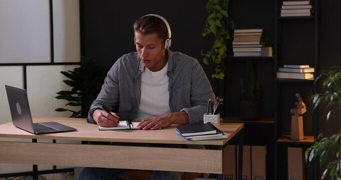 Man in headphones taking notes while passing online test via laptop at table indoors