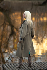 Blonde woman in stylish trench coat stands on wooden dock by tranquil water, with autumn foliage reflecting in the background, showcasing fashion and nature harmony