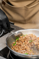 Chef in beige apron and black glove cooking buckwheat with vegetables in a steaming pan on stovetop. Close-up of food preparation in professional kitchen.