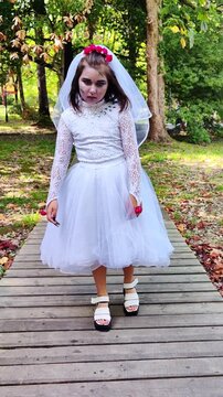 A girl in a white dress and a veil, the image of a scary bride, moves like a zombie on the street, in the park on Halloween