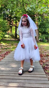 A girl in a white dress and a veil, the image of a scary bride, moves like a zombie on the street, in the park on Halloween